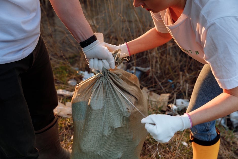 A group of three people, including a man and two children, are engaged in rubbish collection on a sandy beach near a body of water. The man, with dark skin and a beard, is wearing a black cap, red t-s