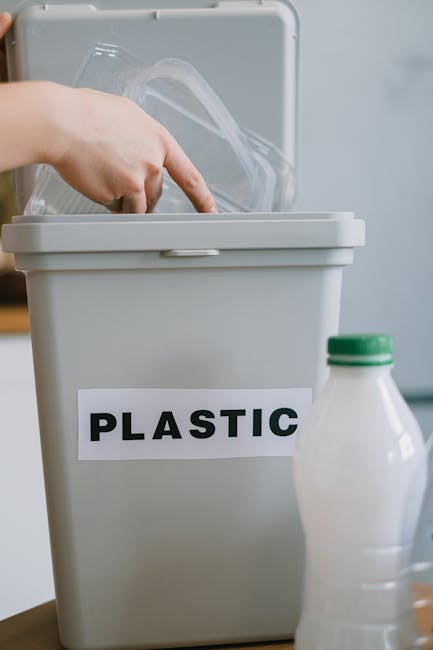 A person's hand is seen placing a transparent plastic container into a grey recycling bin labeled 'PLASTIC' with bold black letters. The bin is positioned on a light-colored surface in an indoor setti