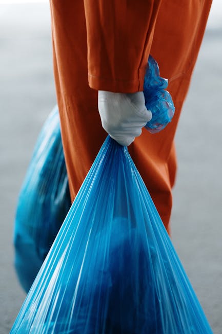 A person's hand is seen placing a transparent plastic container into a grey recycling bin labeled 'PLASTIC' with bold black letters. The bin is positioned on a light-colored surface in an indoor setti